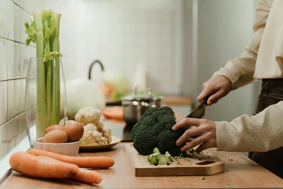 Person chopping broccoli on a wooden board in a kitchen. Surrounding are carrots, eggs, celery, cauliflower, and a pot. Warm lighting.