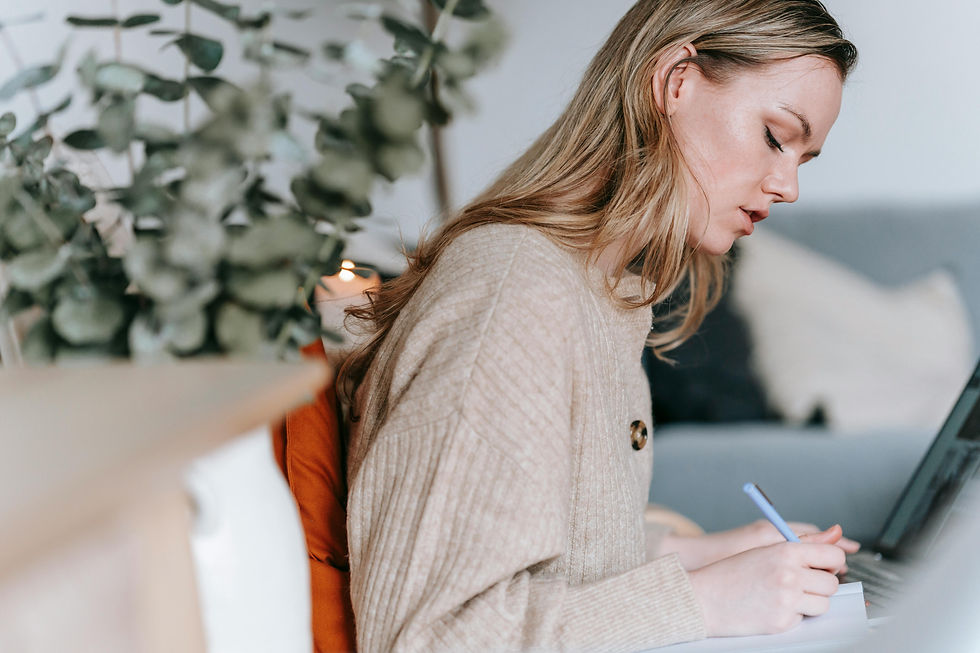 Woman with long hair writes in a notebook, seated on a sofa with plants nearby. Neutral tones create a calm, focused atmosphere.
