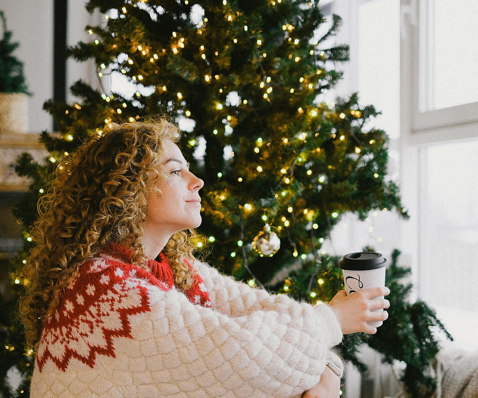 Woman with curly hair holding a coffee cup, sits by a lit Christmas tree, wearing a red and white sweater, gazing out a bright window.