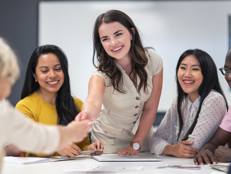 Smiling coworkers in a meeting, one woman in beige shaking hands across a table, with a whiteboard behind them.