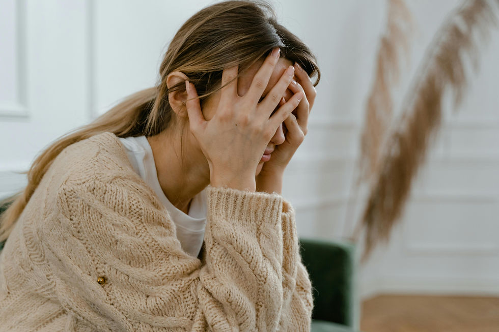 Woman in a beige sweater covers her face with both hands indoors, looking upset or overwhelmed.
