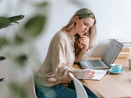 A woman sits at a desk with a laptop and notebook, thoughtfully writing and reflecting, symbolizing intentional planning and evaluating priorities.