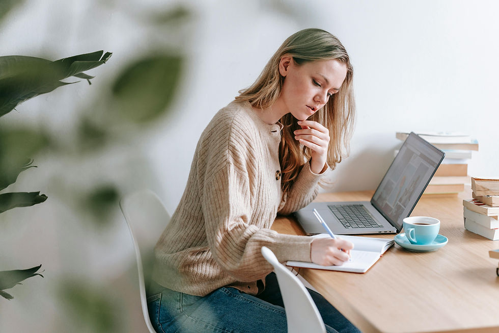 A woman sits at a desk with a laptop and notebook, thoughtfully writing and reflecting, symbolizing intentional planning and evaluating priorities.