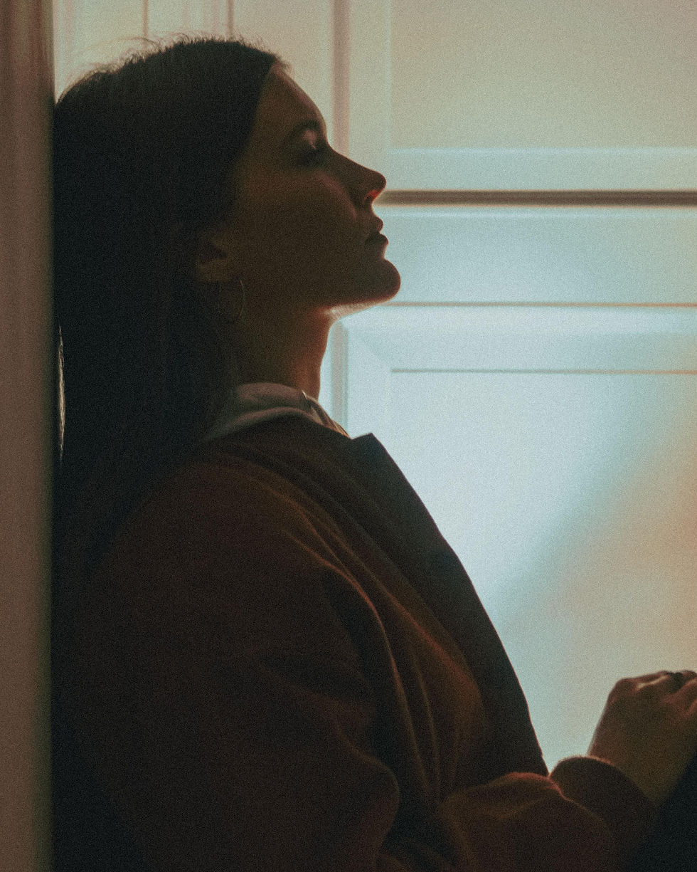 Silhouette of a woman leaning against a wall in a dimly lit room. Soft light creates a contemplative mood. No visible text.