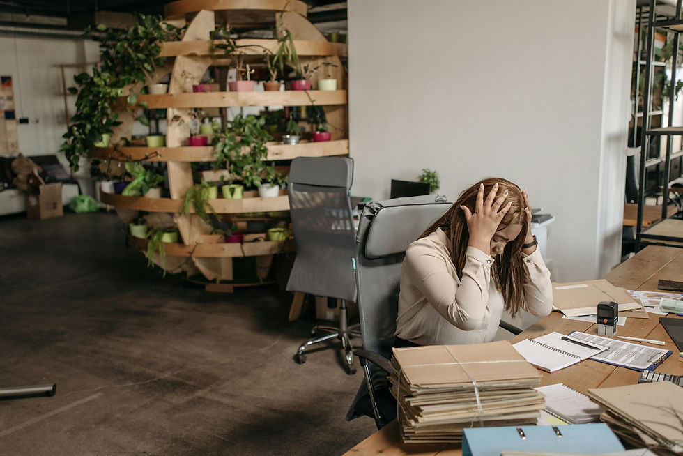 Woman in office holds head in frustration, surrounded by paperwork. Background features plants on wooden shelves. Mood is stressed.