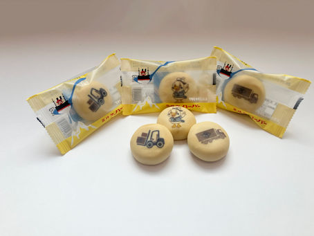 フォークリフトやトラック、キャラクターのイラストがプリントされた丸いお菓子が並んでいる様子。背景には黄色と白の個包装パッケージが配置されており、物流や運送をテーマにしたデザインになっている。