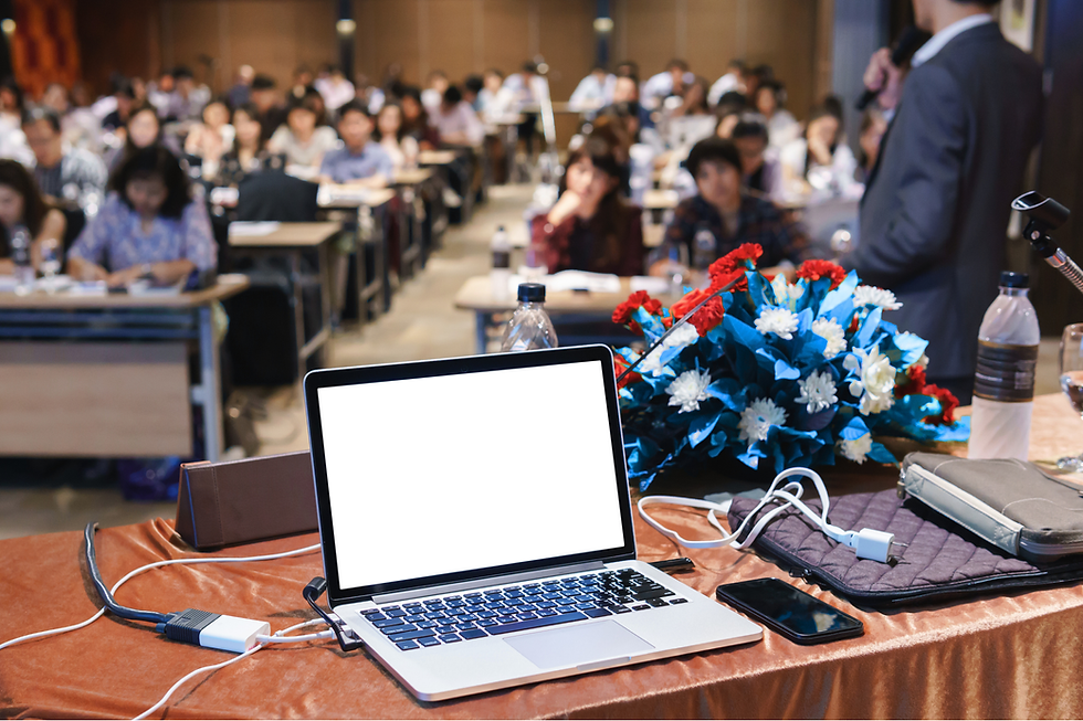 講演会場の壇上から見た風景。手前のテーブルにはノートパソコンやスマートフォン、資料、花束が置かれ、マイクを持つ講師が多くの聴講者に向けて話している様子。
