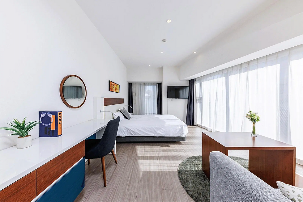 大きな窓から自然光が差し込む、モダンで明るいワンルーム。白い壁と木目調の床、シンプルなベッド、デスクとチェア、壁掛けテレビが配置され、観葉植物や花でナチュラルな雰囲気を演出している。