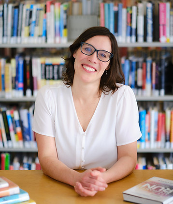 A woman with brown hair and glasses in a white shirt, smiling. There are books in the background.