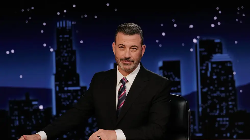 Man in a suit with a colorful tie sits at a desk, smiling slightly. Night cityscape backdrop with starry sky. Studio setting.