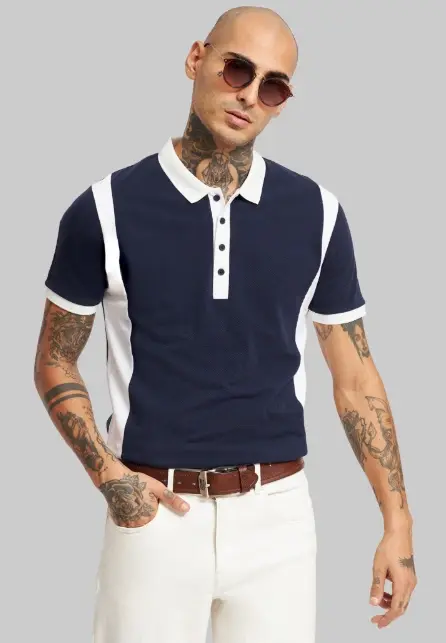 Bald man with tattoos wearing sunglasses, a navy and white polo, and white pants poses against a gray background, exuding a cool vibe.