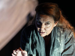 La Rassegna Teatro dei Fabbri de La Contrada, include lo spettacolo con questa donna nell'immagine. Sguardo rivolto verso lo spettatore, leggermente inclinata in avanti indossa uno scialle verde. Il braccio è piegato  ed è visibile la sua mano semiaperta. capelli arancioni, volto in penombra. background nero. Sulla sinistra una forma beije.