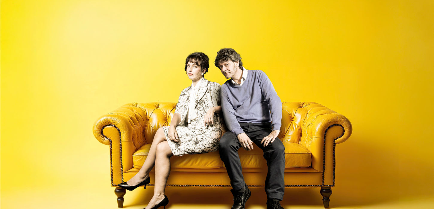 Marzia Postogna e Maurizio Repetto per lo Spettacolo La Felicità. Entrambi seduti su un divano giallo trapuntato. Lei ha le gambe acavallate e indossa un vestitino. Background giallo. Uno spettacolo che andrà in scena al Teatro Bobbio de La Contrada di Trieste.