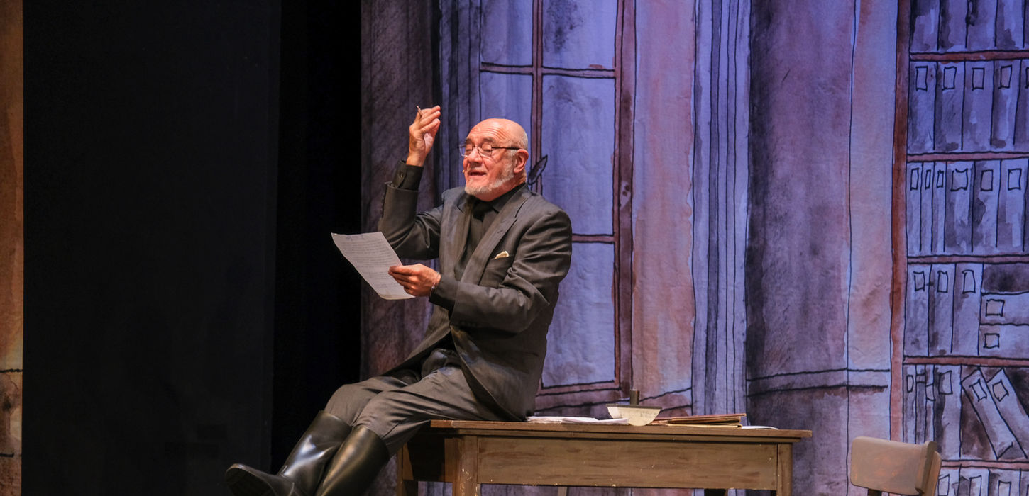 nella foto Maurizio Zacchigna è seduto su un tavolo e ha le gambe incrociate, tiene in mano un foglio di carta che legge con aria un po' beffarda. Sullo sfondo ci sono le scenografie di Andrea Stanisci. uno spettacolo che andrà in scena al Teatro Bobbio de La Contrada di Trieste