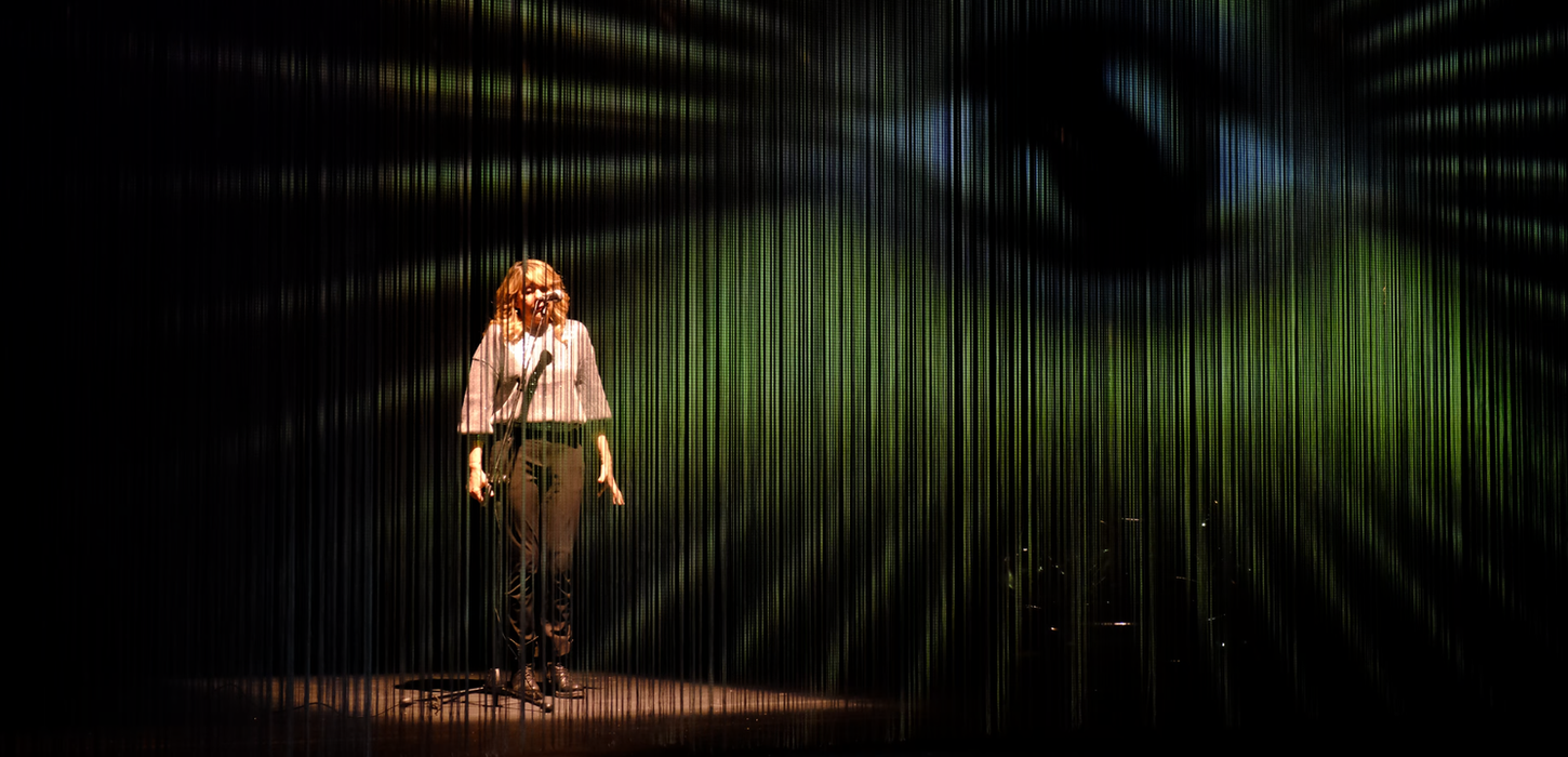 immagine con la foto di Emanuela Grimalda in scena, con una proiezione di un occhio e una piramide in giallo. Lo spettacolo è una produzione Teatro Bobbio de La Contrada Teatro Stabile di Trieste