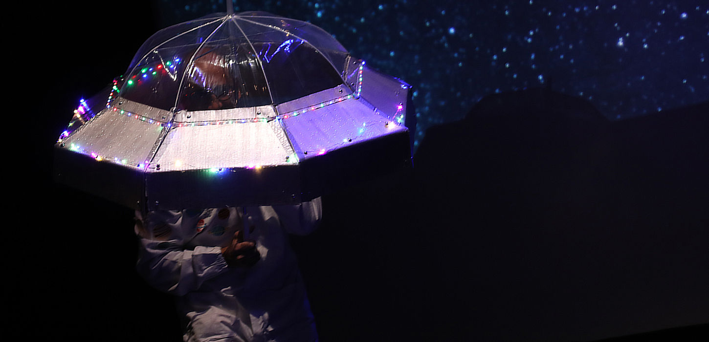 Foto di Valentino Pagliei travestito da astronauta, con un ombrello a forma di navicella spaziale. lo sfondo è un cielo stellato. Lo spettacolo andrà in scena al Teatro Bobbio de La Contrada Teatro Stabile di Trieste