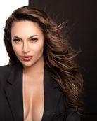 A headshot of a female model on location in Oahu with long, wavy brown hair. She wears a black blazer against a split white and black background, looking directly at the camera with a confident expression.
