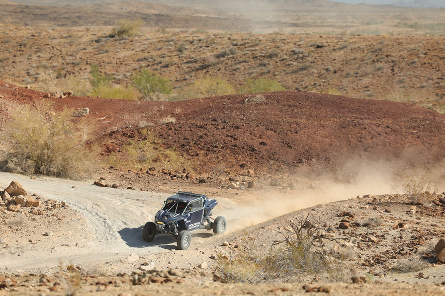Black quad dirt racing car speeding through a desert track surrounded by hills of desert brush.