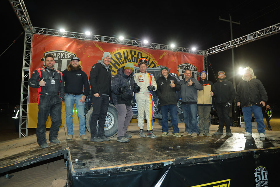 Mike Mamelli and friends posing infront of his black quad car on racing podium at night.
