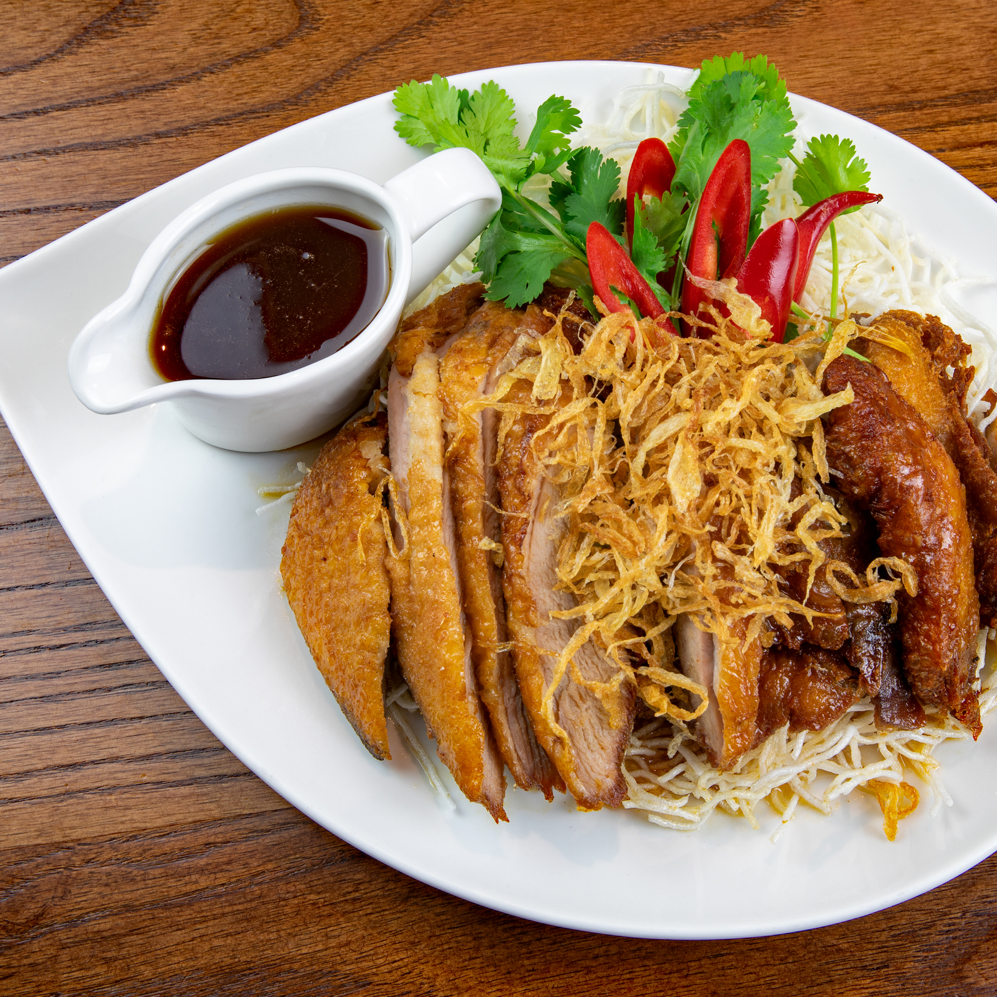 Red Elephant Siam, Thai restaurant, East Dean, Eastbourne. Signature dish. Slices of duck garnished with crispy shallots, served with a gravy boat of tamarind sauce on a white ceramic teardrop-shaped plate placed on a wooden table.