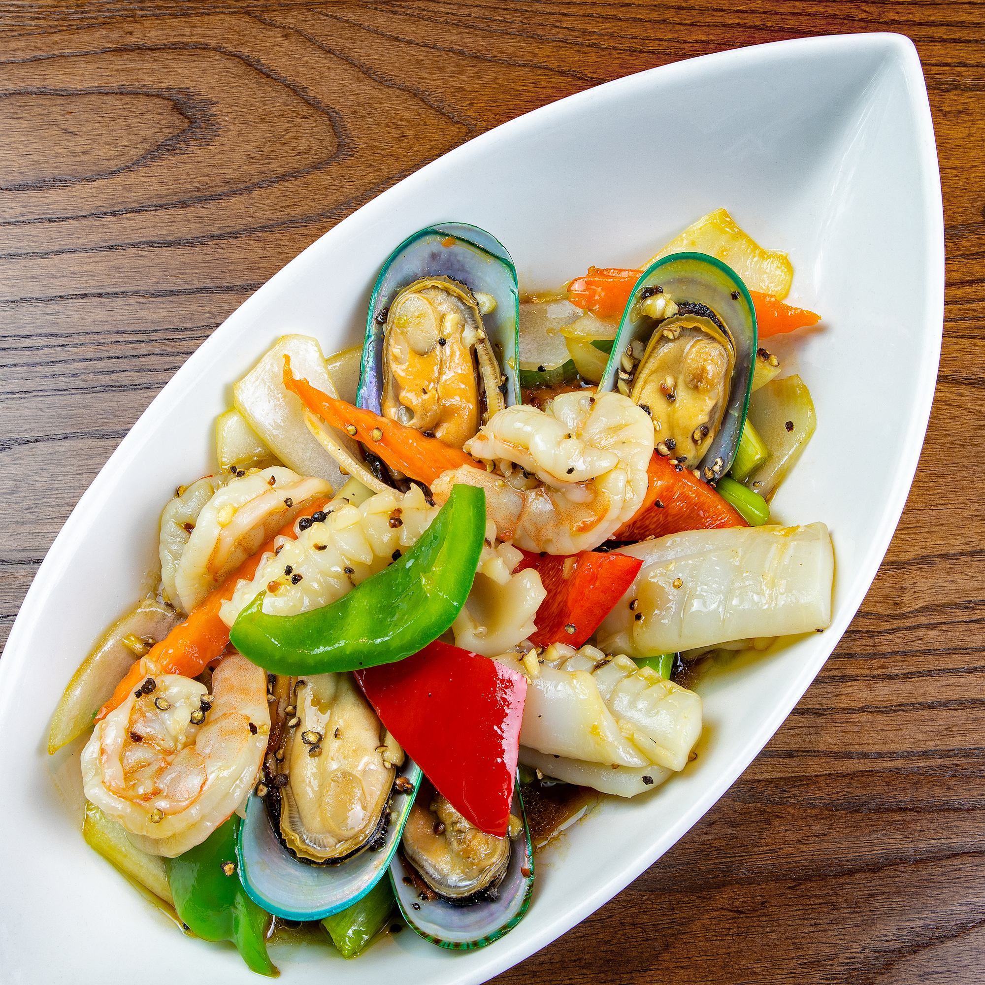 Red Elephant Siam, Thai restaurant, East Dean, Eastbourne. Signature dish. Stir fried Garlic/Kratiem Prik Thai with mixed seafood, served in a white ceramic boat-shaped bowl placed on a wooden table.