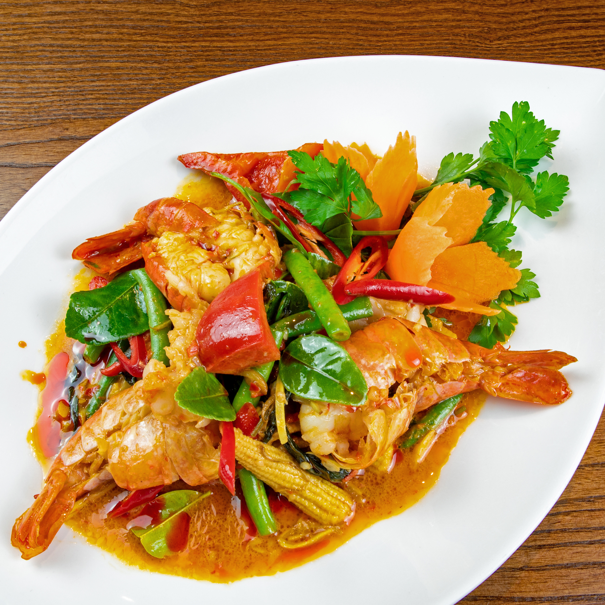 Red Elephant Siam, Thai restaurant, East Dean, Eastbourne. Signature dish. Giant Prawn Pad Cha garnished with sweet peppers, baby corns, chillies, lime leaves and an orange spicy sauce, served on a white ceramic teardrop-shaped plate placed on a wooden table.