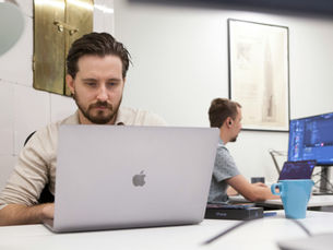 Two men work intently on laptops in a modern office. A blue mug and iPhone box are on the desk. A framed city sketch hangs on the wall.
