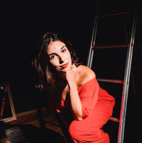 Gabi Magnani é uma mulher branca de cabelos e olhos castanhos. Gabi está vestida toda de vermelho com pés descalços. Uma de suas mãos está no seu queixo e ela está sentada em uma escada, nos bastidores de um teatro. A luz é cênica e um com sombras.