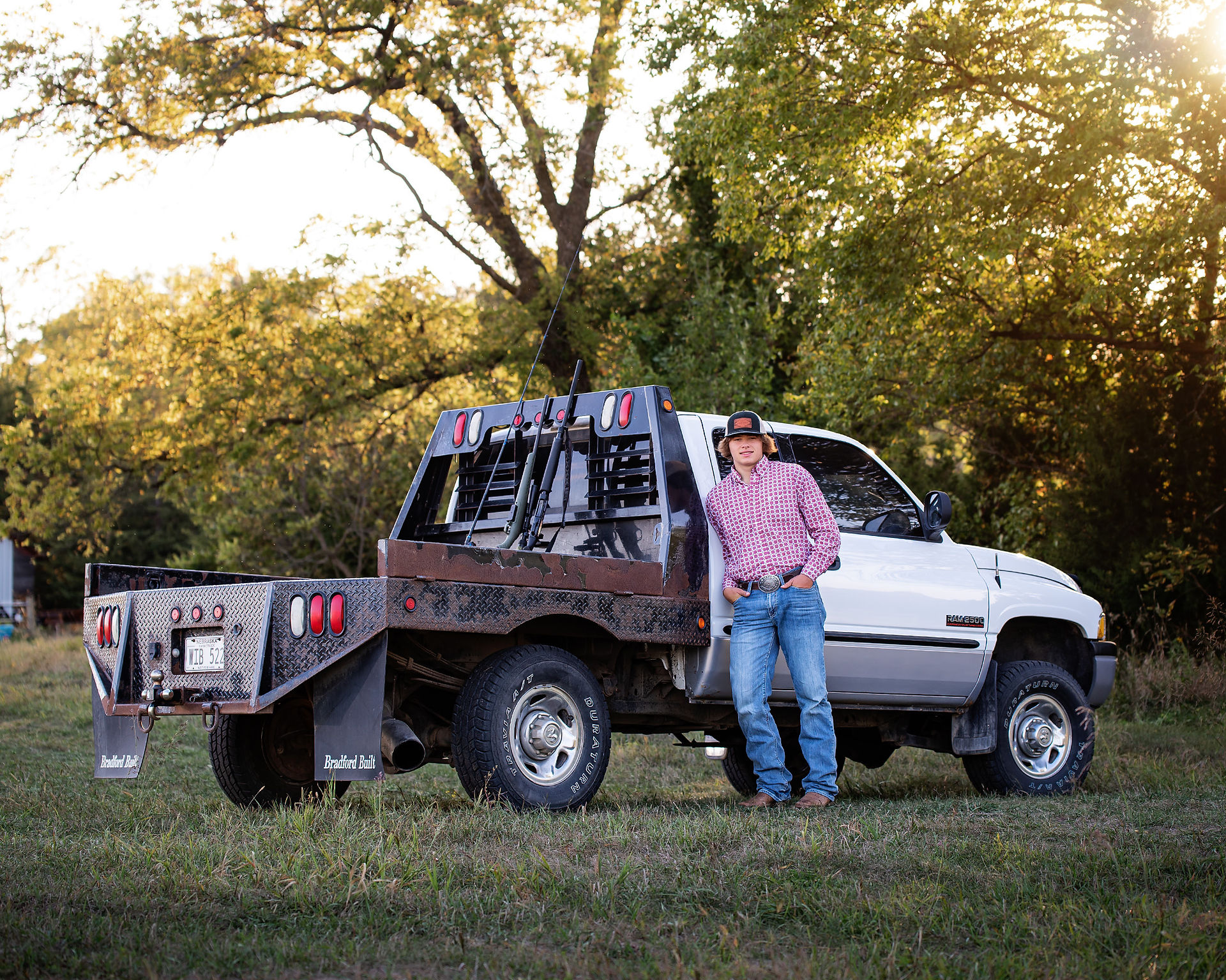 A boy leaning against pick up truck with hunting rifles and fishing pole mounted in the back for senior pictures.