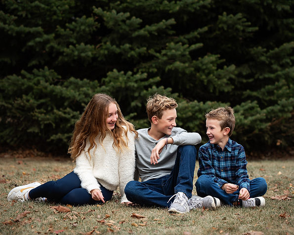 Three siblings laughing at each other while sitting on the ground during family pictures