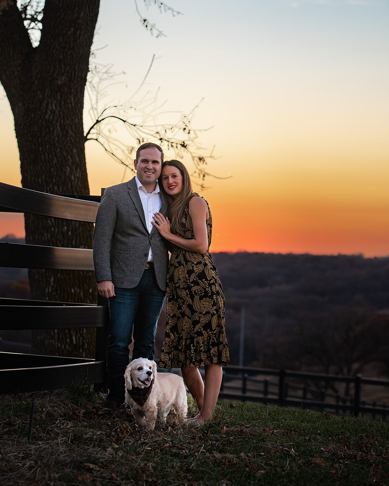 Husband and wife posing next to wooden fencing with family dog during an autumn sunset in Omaha, Nebraska.