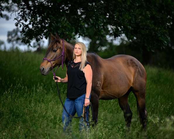 A woman stands next to her horse in a grassy pasture, with a powerful stoic smile.