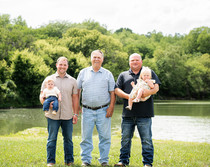 Grandfather standing with his two sons holding their sons in an open field in front of of a lake for extended family gathering event.