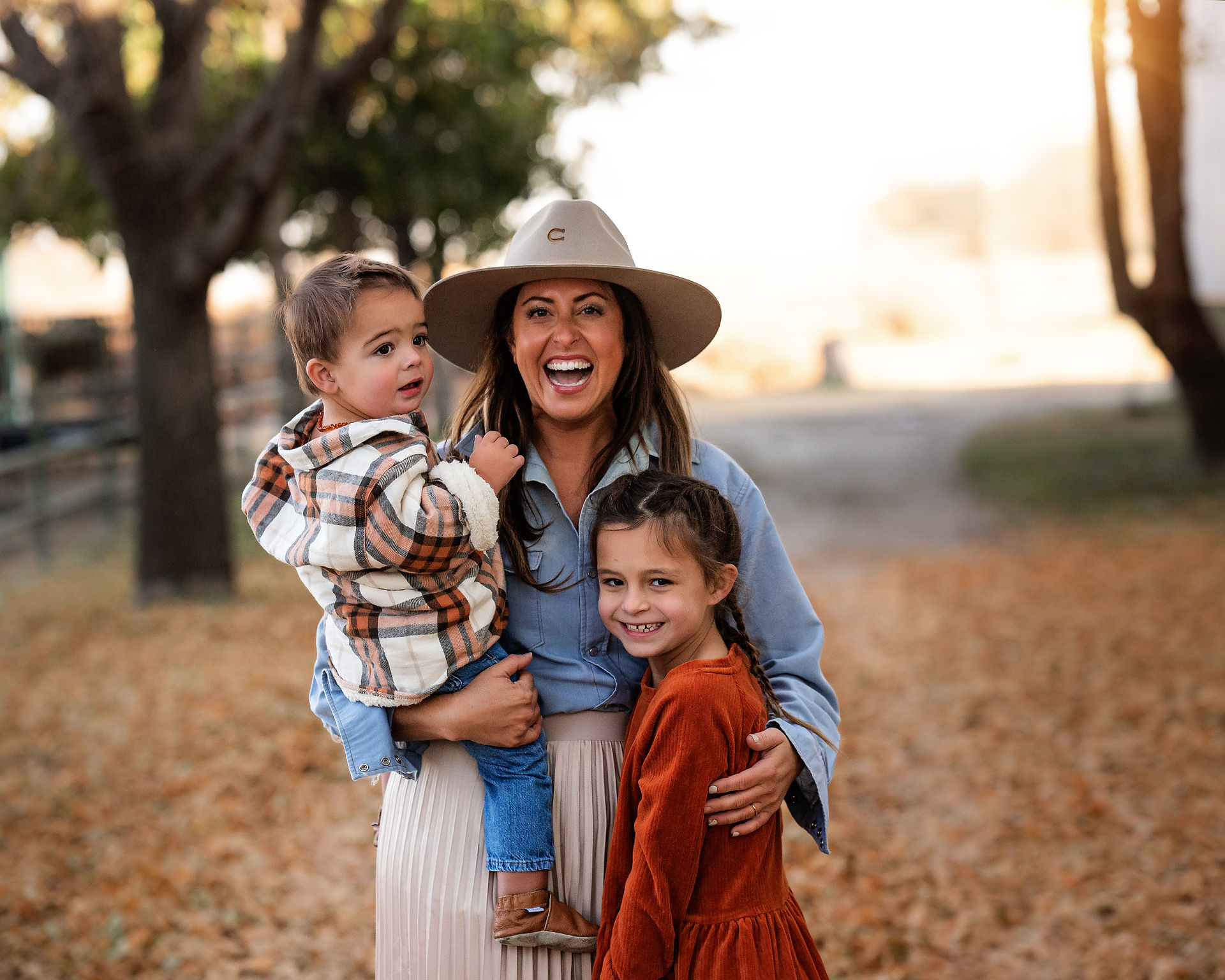 Mother holding youngest child and hugging daughter with beautiful fall scenery with autumn leaves covering the ground in Ashland, Nebraska.