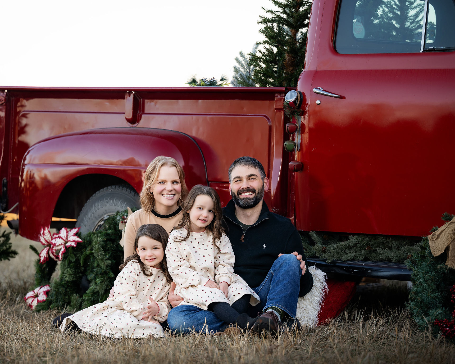 A family with young children nestle next to a vintage red pickup truck for a holiday photo.