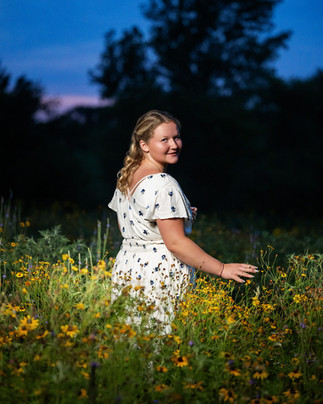 Senior girl in a white floral dress reaches into a wildflower patch at dusk in Eagle, Nebraska