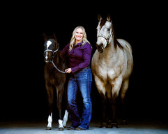 Woman standing with horses in stable walkway with black backdrop to bring out the contrast of coat colors. 