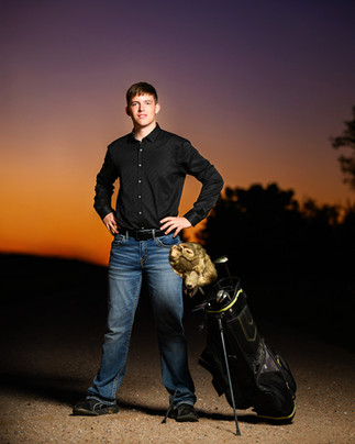 Senior guy stands on a dirt road at dusk with his golf bag beside him under a purple and orange sky in McCook, Nebraska