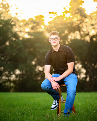 Senior guy in a black polo and jeans sits on a wooden stool against a golden treeline in Gretna, Nebraska