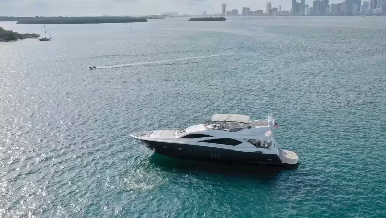 A sleek black and white luxury motor yacht floating on vibrant turquoise water with the Miami skyline visible in the background under a partly cloudy sky.