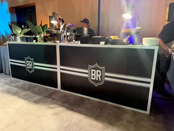 Sleek black branded catering station with double white racing stripes and the BR shield logo, staffed by chefs working under metal heat lamps.