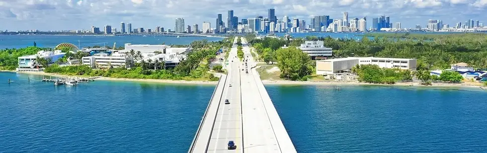 A panoramic view of a long, straight road extending over the water, leading toward a city skyline in the background. The scene includes greenery on both sides of the road, with buildings on the shore and water glistening under clear skies.