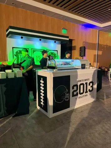 Custom food station with football laces detailing, a helmet icon, and 'Born to Compete 2013' branding, backed by framed jerseys displayed under green mood lighting.
