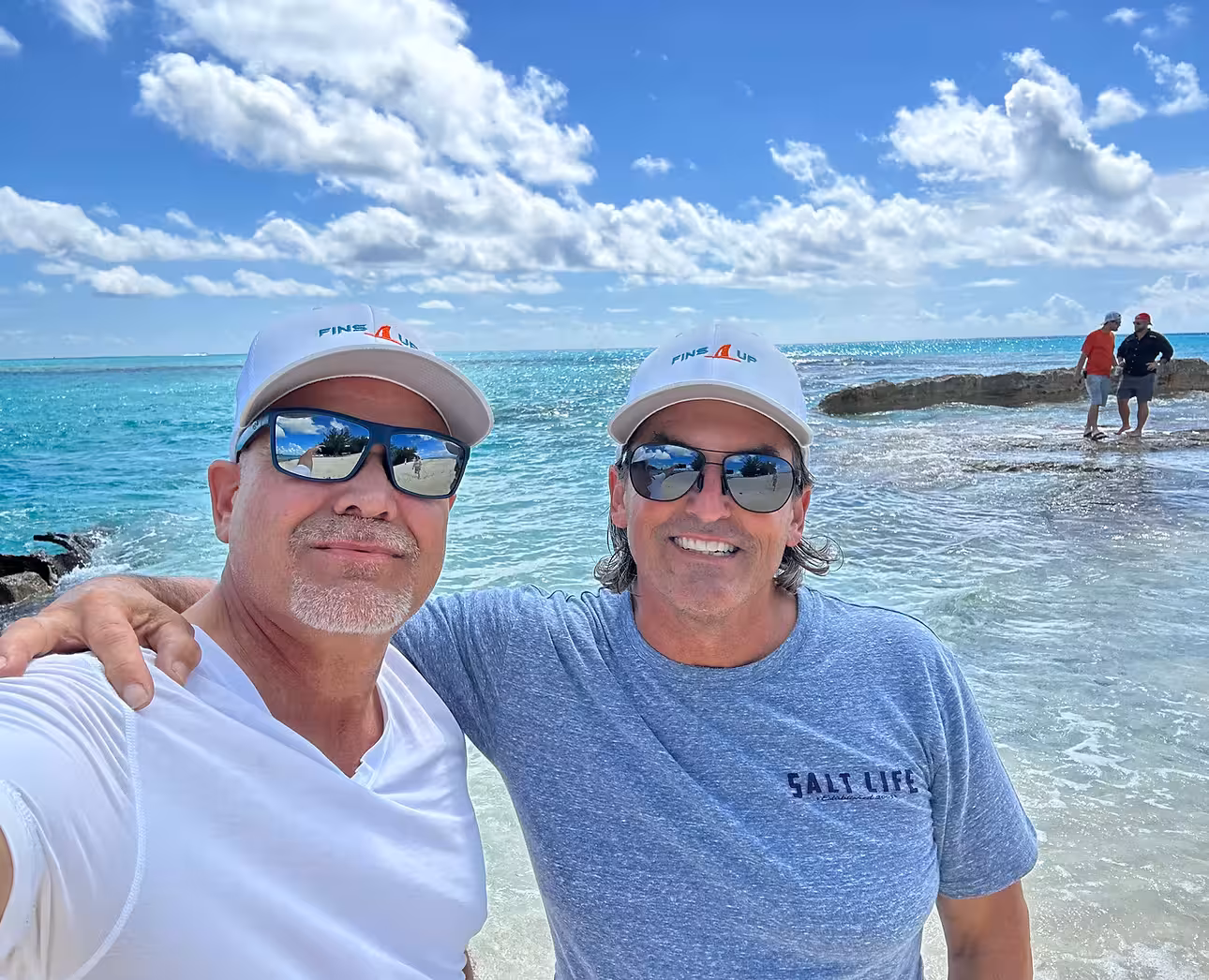 Selfie of two men smiling on a sunny tropical beach. Both are wearing white 'FINS UP' caps and sunglasses. The man on the right wears a blue Salt Life shirt and has his arm around the man on the left, who is wearing a white shirt. Behind them is clear turquoise water, a rocky shoreline, and two people standing in the distance under a bright blue sky with scattered clouds.