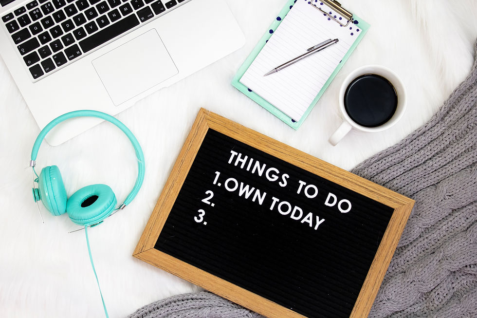 A laptop, headphones and notebook lie on a table alongside a cup of a coffee and the words "things to do: Own today" on a small blackboard