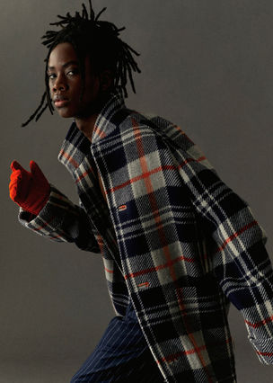 Young male model wearing plaid coat, orange gloves, and striped pants.