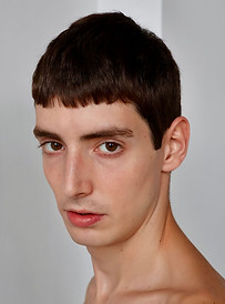 Young man posing shirtless with short brown hair against a gray background.