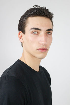 Mario's confident gaze complements his relaxed, open-collar white shirt and chic necklace, against a serene seascape background at sunset, exuding effortless Mediterranean elegance.