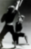 Two people engaged in a martial arts duel with sticks. One kneels, striking upwards, the other lunges. Black and white, intense focus.