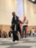A martial artist in black attire with the USA flag trains on a mat. Onlookers record with phones. "WAKO USA" is projected on the wall.